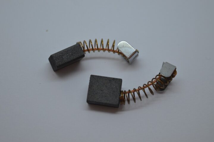 Two carbon motor brushes with coiled copper springs laid on a white surface, shown in a curved arrangement.